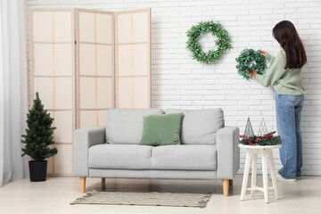 Young woman hanging Christmas wreath on white brick wall in living room