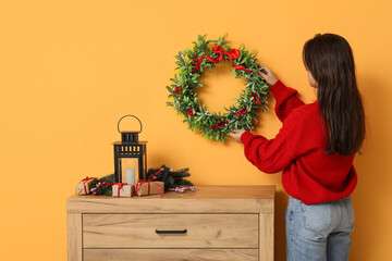 Young woman hanging Christmas wreath on yellow wall in living room