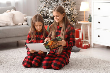 Cute happy little girls in checkered pajamas with book reading Christmas story at home