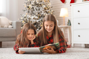 Cute happy little girls in checkered pajamas with book reading Christmas story at home