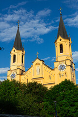 Fototapeta premium View of Church of Assumption of Mary with statue of Virgin Mary in niche on main facade of Romanesque building and two steeples rising above green trees against blue sky in Italian town of Bruneck