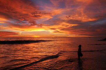Fototapeta premium A silhouette of a person stands in the shallow water, creating a sense of solitude and reflection. The gentle waves lap at the shore, mirroring the colors of the sky. 