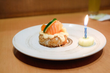 A slice of cucumber and fresh salmon on top of rice cake and a burning candle on white plate