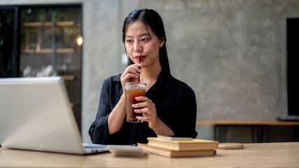 A beautiful Asian businesswoman enjoys iced coffee while working on her laptop, working remotely.