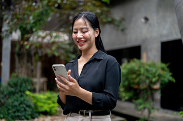 A charming Asian businesswoman is reading text messages on her smartphone in a garden space.