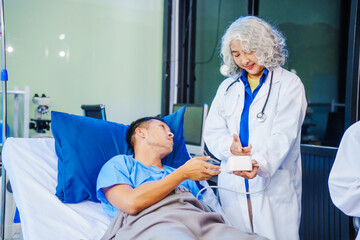 Obraz premium A middle-aged man lies bedridden on hospital bed, closely monitored by an old Asian woman doctor and young nurse. They assess his symptoms, including heart and lung conditions, offer advice.