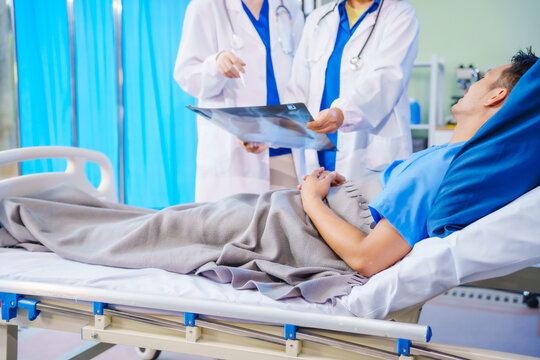 A middle-aged man lies bedridden on hospital bed, closely monitored by an old Asian woman doctor and young nurse. They assess his symptoms, including heart and lung conditions, offer advice.