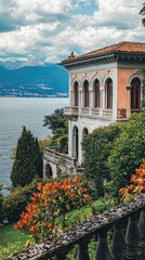 Naklejka premium Charming lakeside villa surrounded by lush greenery and vibrant flowers, with stunning mountain views under a partly cloudy sky.