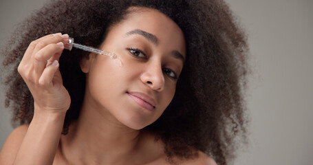 Obraz premium Beauty and healthcare concept - beautiful african american woman with curly afro hairstyle and clean, healthy skin applies skin care serum from a pipette to her skin