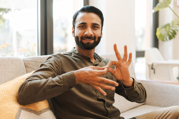 Smiling indian man is sitting on sofa and showing sign language gesture looking at camera