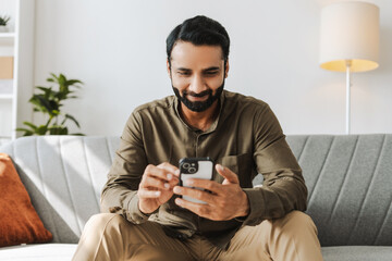 Smiling Indian man sitting on sofa holding mobile phone, using mobile app at home, text message