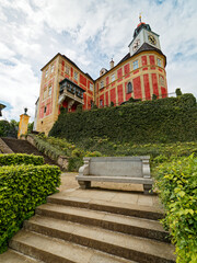 Castle Jánský Vrch is situated in the town of Javorník in north Moravia on the border with Poland. This image shows the castle's red facade, a clock tower, ivy-covered walls, and a stone bench below.
