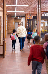 Fototapeta premium Walking in school hallway, female teacher guiding young diverse students with backpacks