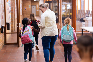 Walking in school hallway, female teacher guiding diverse students with backpacks to classroom