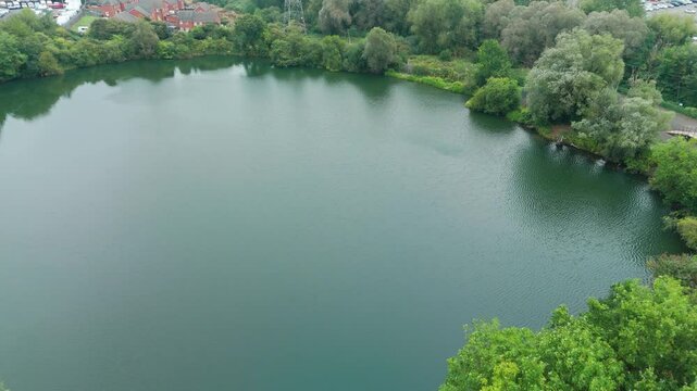 Aerial shot of a serene lake bordered by lush trees and adjacent to a residential area, showcasing a picturesque blend of nature and human habitation. The calm water reflects the greenery and rooftops