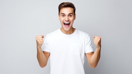 Naklejka premium A joyful man in a white t-shirt clenches his fists in excitement against a plain backdrop, capturing a moment of triumph and exuberance.