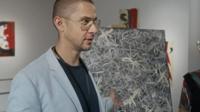 Medium close-up of handsome young Caucasian male art critic talking to public about new contemporary art painting, displayed on easel at exhibition in gallery, during champagne party at opening