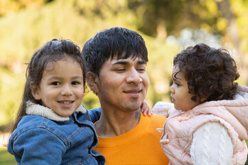 Headshot of happy hispanic father holding his children outdoors while his son is looking at the...