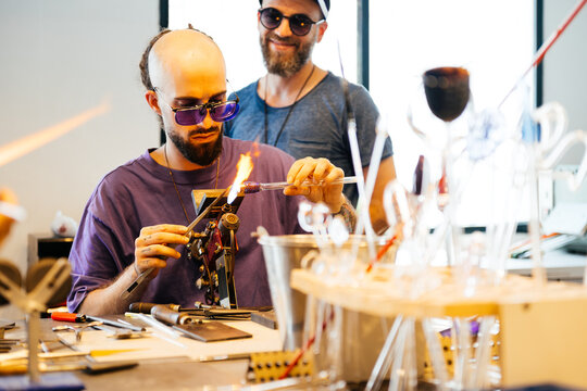 A glassblowing artisan concentrates on heating a glass tube with a torch as they create a piece in their studio.