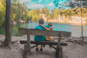 Two brothers sitting on wooden deck chair in the rays of summer sun near lake in rocks Adrspach in the Czech republic. Blue sky, forest and lake. Summer concept.