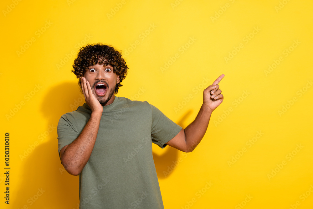 Poster Photo of astonished guy with curly hairstyle dressed khaki shirt indicating at promotion empty space isolated on yellow color background