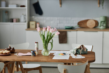 Fresh tulips in vase on wooden table surrounded by various crafting supplies, including paper hearts, markers, and scissors in cozy kitchen setting