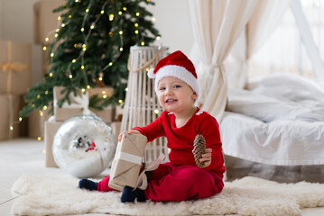 Smiling little baby boy in a santa costume and red hat sitting on a soft fur carpet in front of a christmas tree