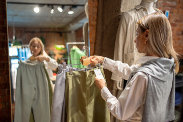 Interested middle aged woman shopping for eco outfit, holding hanger of natural organic cotton...