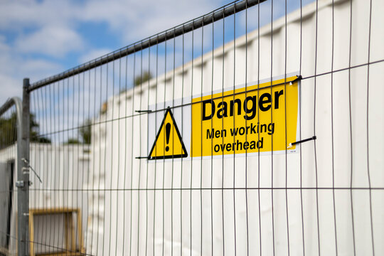 Yellow and black Danger Men Working Overhead triangle warning sign on construction site temporary heras fence panel and white steel storage container