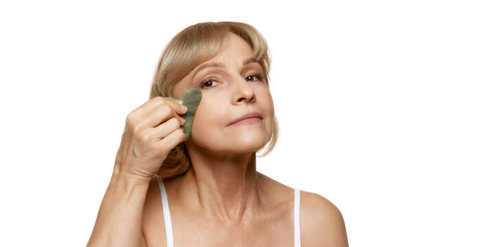 Woman uses scrapper under her eye, enhancing her skincare routine with natural beauty tools against white studio background. Concept of beauty, wellness, cosmetology, anti-aging products. Ad