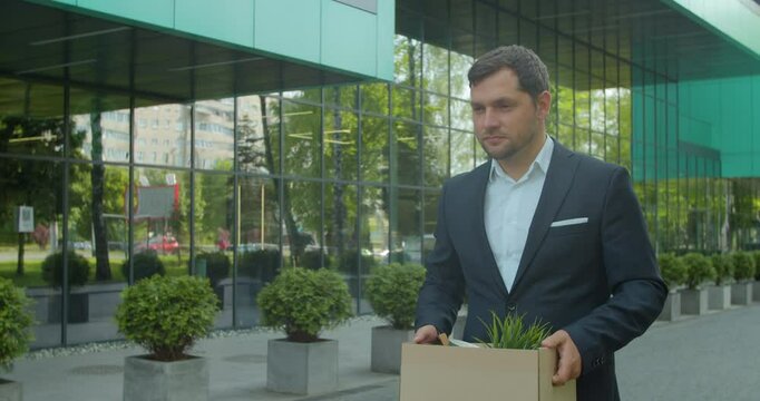 Businessman gets fired and walking near office center with carton box and documents with a table flower. Lost His Job. Unemployment rate growing due pandemic.
