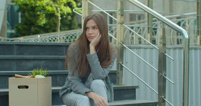 Portrait of young workless woman siting on stairs near office center with carton box and documents with a table flower Need job. Female fired office worker in depression. Unemployment rate growing due