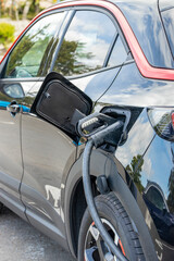 Black electric vehicle (EV) connected by cable to public plug-in charging station in car park