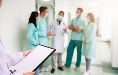 Fototapeta premium A group of masked healthcare professionals in lab coats and scrubs discussing medical matters in a hospital hallway, with a close-up of a clipboard in the foreground. Teamwork and healthcare themes.