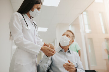 A female doctor, wearing a mask and a stethoscope, gently holds a male patient's hand in a hospital corridor. Both wear masks, emphasizing care, compassion, and safety in a medical setting.