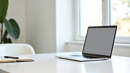 A modern workspace featuring a laptop, notebook, and stationary on a white desk with greenery in a bright room