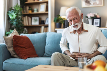 Senior man suffering stomach ache sitting on a couch in the living room, Mature man holding his...
