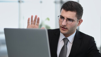 A young man in a black suit makes a welcome gesture on the webcam.