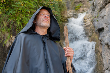 A monk in a black cassock with a wooden staff against the background of a mountain waterfall.