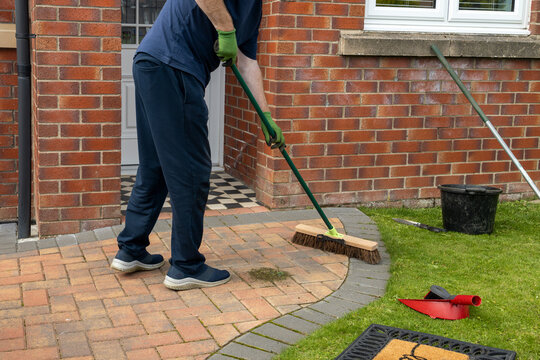 Man sweeping a brick driveway path in front of a house with bucket, broom, and other cleaning tools nearby