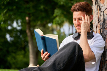 A man is sitting on a tree and reading a book. He is wearing headphones and has a smile on his face
