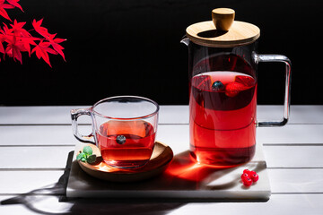 Red fruit infusion in crystal glass and pitcher on white wooden table on dark background.