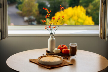 Cozy autumn table setting with pumpkin pie, coffee cup, orange flowers in vase, and a small pumpkins by a window with yellow foliage. Autumn, fall, Thanksgiving concept.
