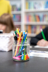 Glass jar filled with pencils sits on table during drawing lesson in library. In blurred...