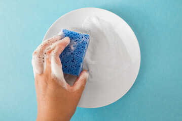 A hand holds a plate with a cleaning sponge with soap foam on a blue isolation background