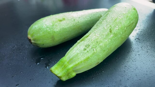 Two fresh zucchini with drops of water lie on a black kitchen table and the camera spins around them