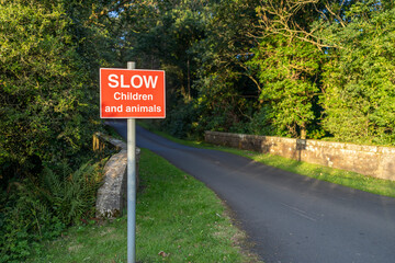 Red road sign warning drivers to slow down due to children and animals on a narrow stone bridge and road lined with trees