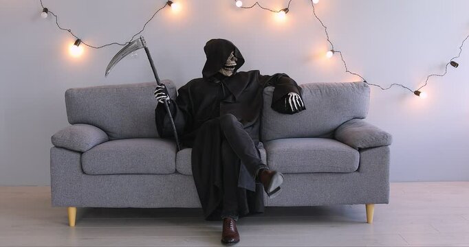 Funny man in Halloween grim reaper death costume holding scythe in black cloak sitting on sofa on gray background with festive garland. Halloween party and horror concept. 4k video.