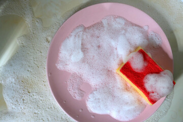 Red sponge for washing dishes with a pink plate in soap foam on the kitchen table top view