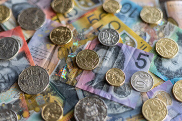 Close-up from above of multiple Australian dollar banknotes and coins spread over a desk, with sharp focus on a one Australian dollar coin, over a 5 dollar banknote and blurred surroundings.